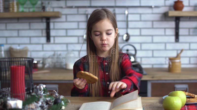 Menina bonita pequena para sentar-se na mesa de cozinha, comendo cookies e folheando através do livro