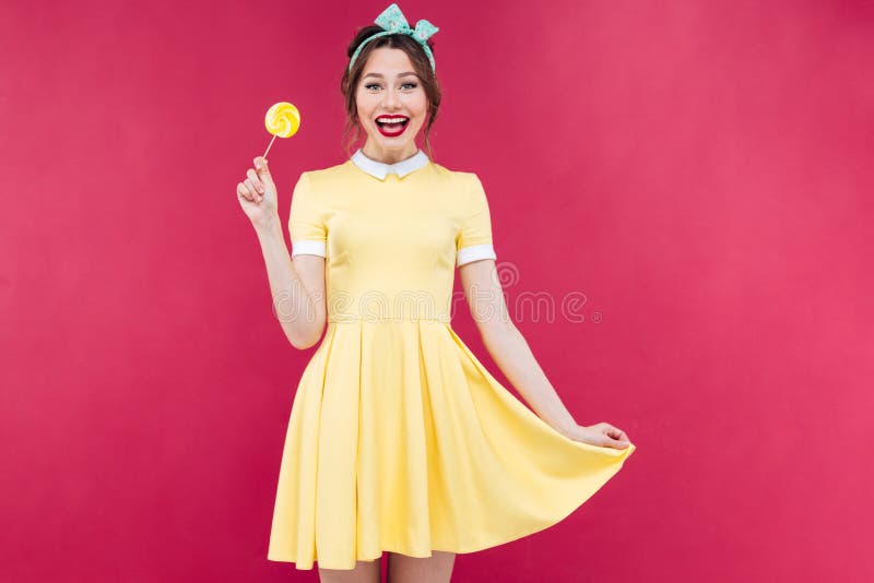Menina bonito alegre do pinup que está e que guarda o pirulito amarelo fotografia de stock royalty free Menina bonito alegre do pinup que está e que guarda o pirulito amarelo fotografia de stock royalty free