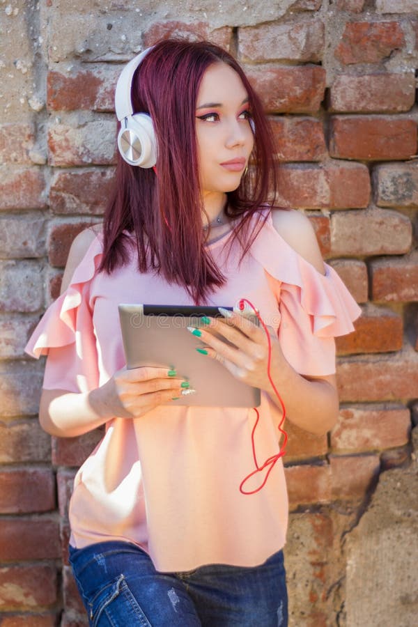 Menina bonito do ruivo que escuta a música na tabuleta digital com os fones de ouvido contra a parede de tijolo exterior imagens de stock royalty free Menina bonito do ruivo que escuta a música na tabuleta digital com os fones de ouvido contra a parede de tijolo exterior imagens de stock royalty free