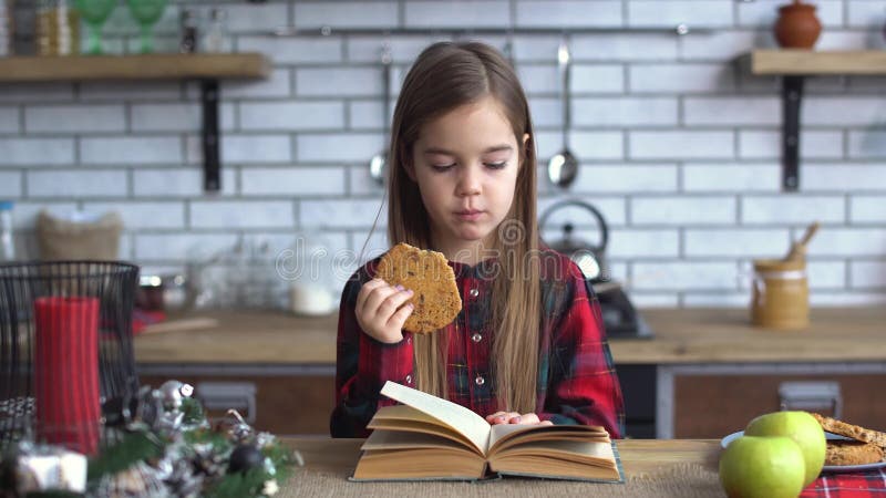 Menina bonito em uma camisa de manta e com cabelo longo que come cookies e que folheia através de um livro na mesa de cozinha