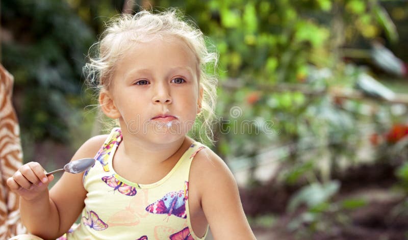 A menina bonito lambe uma colher com um gelado delicioso na A.A. fotografia de stock royalty free A menina bonito lambe uma colher com um gelado delicioso na A.A. fotografia de stock royalty free