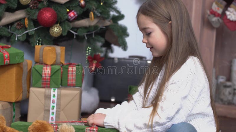 Menina bonito pequena com o cabelo marrom longo que senta-se perto da árvore de Natal e dos presentes abertos