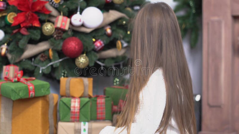 Menina bonito pequena com o cabelo marrom longo que senta-se perto da árvore de Natal e dos presentes abertos