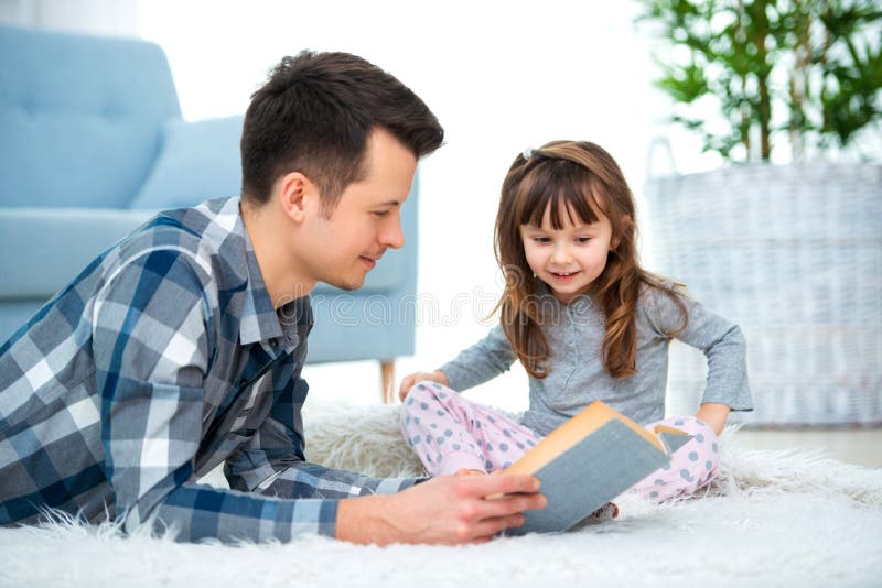 Menina bonito que escuta o conto de fadas da leitura do paizinho que encontra-se no assoalho morno junto, importando-se o livro d fotografia de stock Menina bonito que escuta o conto de fadas da leitura do paizinho que encontra-se no assoalho morno junto, importando-se o livro d fotografia de stock