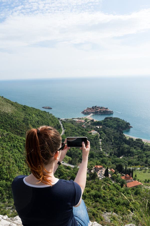 Menina branca nova que faz a tomada de uma foto com o telefone esperto de Sveti Stefan em Budva, Montenegro Penhasco verde e pais imagem de stock royalty free Menina branca nova que faz a tomada de uma foto com o telefone esperto de Sveti Stefan em Budva, Montenegro Penhasco verde e pais imagem de stock royalty free