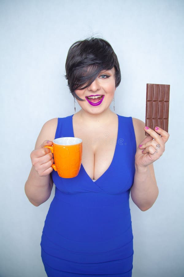 Menina carnudo bonito com cabelo preto curto em um vestido azul que guarda uma barra de chocolate e um copo em suas mãos, sorrind fotografia de stock royalty free Menina carnudo bonito com cabelo preto curto em um vestido azul que guarda uma barra de chocolate e um copo em suas mãos, sorrind fotografia de stock royalty free