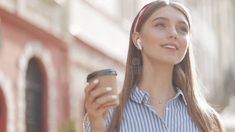 Menina caucasiana muito feliz com cabelo castanho e camisa com pulseira em fones de ouvido sorrindo bebendo