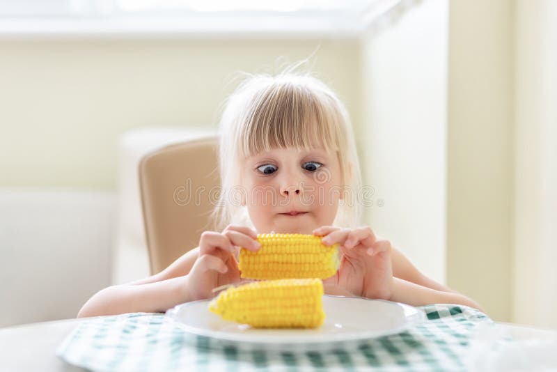 Cute blond caucasian girl holding in hand tasty boiled sweet corn cob and lokking on it with amazed wide opened eyes. Child loves eating  yellow maize. Children summer snack kid food healthy vegetable childhood cheerful young lunch hungry portrait happy adorable bite organic fun toddler female natural meal looking happiness indoor kitchen home window amazing adore like crazy. Cute blond caucasian girl holding in hand tasty boiled sweet corn cob and lokking on it with amazed wide opened eyes. Child loves eating  yellow maize. Children summer snack kid food healthy vegetable childhood cheerful young lunch hungry portrait happy adorable bite organic fun toddler female natural meal looking happiness indoor kitchen home window amazing adore like crazy