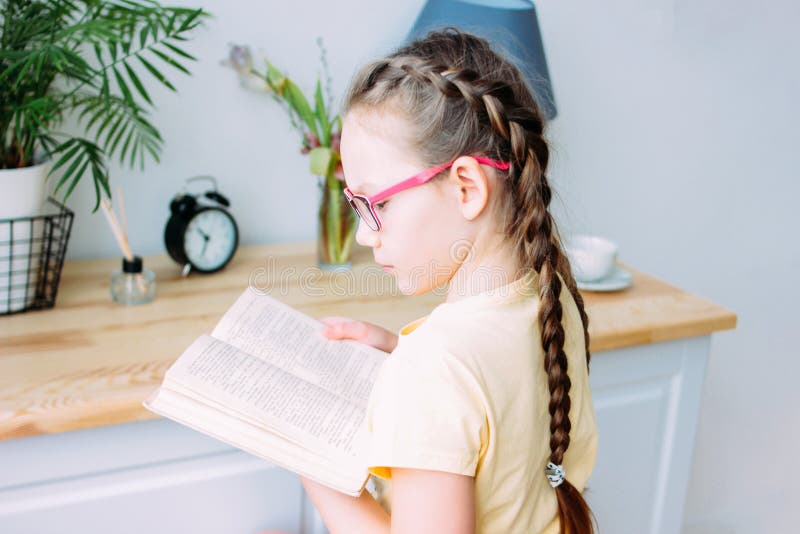 Menina com o livro de leitura dos vidros e das tranças na tabela em sua sala fotografia de stock Menina com o livro de leitura dos vidros e das tranças na tabela em sua sala fotografia de stock