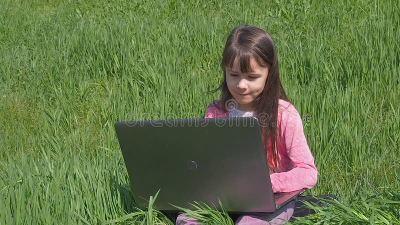 Menina com o portátil na grama verde Menina na natureza Uma criança bonita com um portátil é contratada em um parque