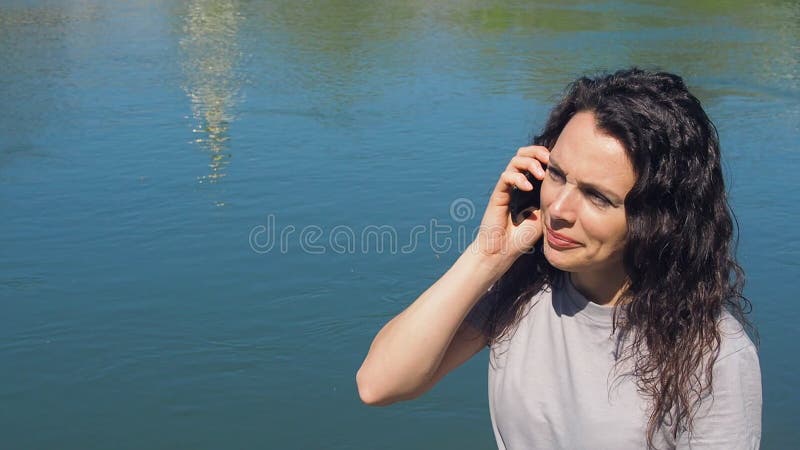 Menina com telefone celular pela água Uma mulher está falando no telefone na terraplenagem do rio em um dia ensolarado