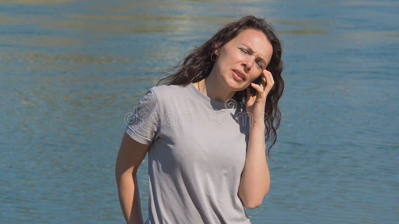 Menina com telefone celular pela água Uma mulher está falando no telefone na terraplenagem do rio em um dia ensolarado