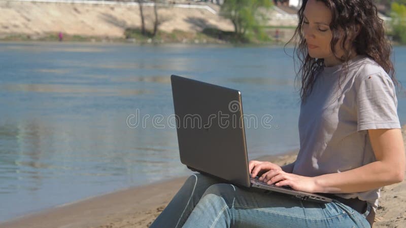 Menina com um portátil pela água Trabalhe no computador no ar fresco pelo rio Dia ensolarado da mola na natureza