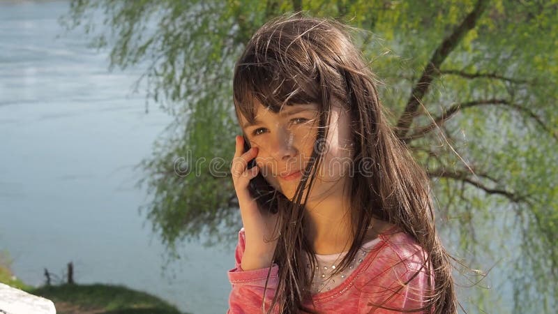 Menina com um telefone pela água A criança está no ar livre Uma menina em um dia de mola ensolarado pelo rio