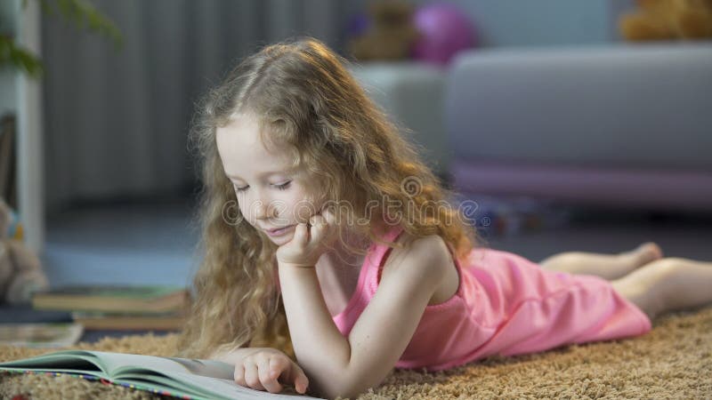 Menina curiosa que encontra-se no assoalho, lendo o livro de histórias das crianças interessantes