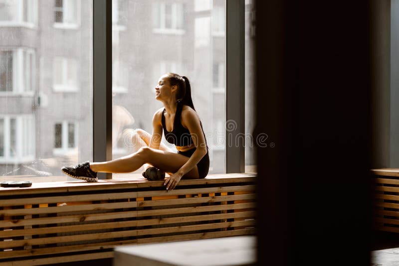 A menina de cabelo escuro atl?tica vestida na parte superior e no short pretos dos esportes est? sentando-se em um peitoril de ma fotografia de stock A menina de cabelo escuro atl?tica vestida na parte superior e no short pretos dos esportes est? sentando-se em um peitoril de ma fotografia de stock