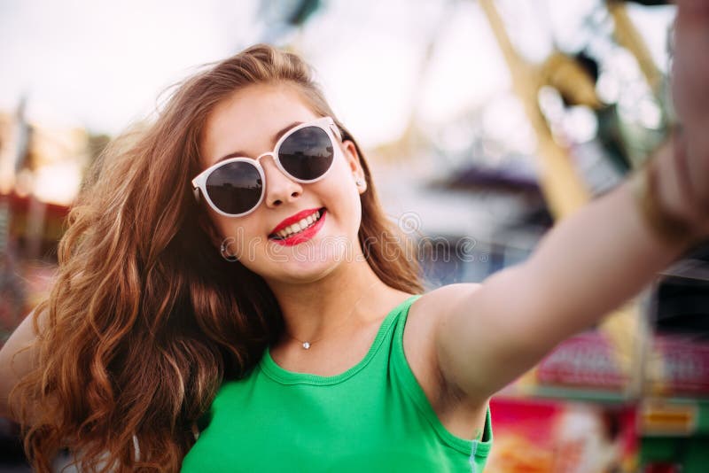 Menina de sorriso alegre nos óculos de sol cor-de-rosa que fazem o selfie na frente do carrossel Retrato exterior da mulher à mod imagem de stock royalty free Menina de sorriso alegre nos óculos de sol cor-de-rosa que fazem o selfie na frente do carrossel Retrato exterior da mulher à mod imagem de stock royalty free
