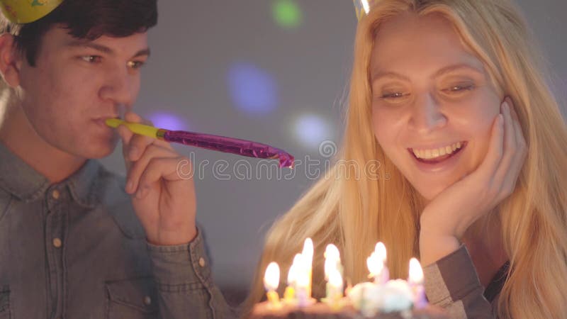 A menina de sorriso loura e um homem nos chapéus do aniversário que sentam-se na frente de pouco bolo com velas A mulher tem o an