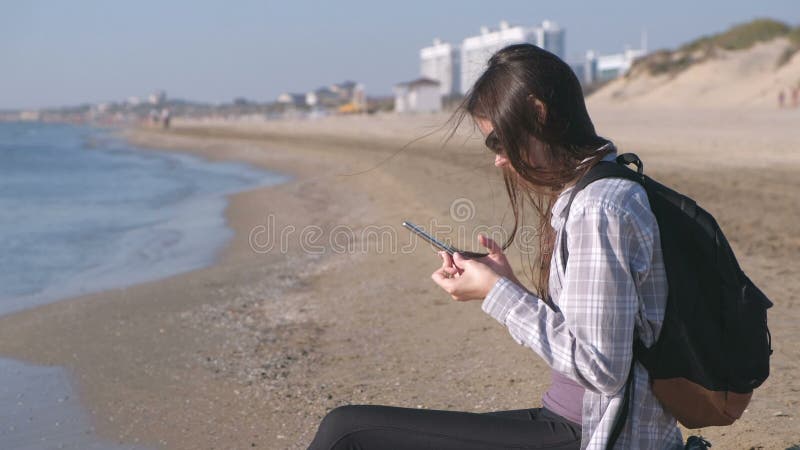 Menina do viajante com uma trouxa que senta-se em uma praia arenosa do mar e que datilografa uma mensagem no telefone celular