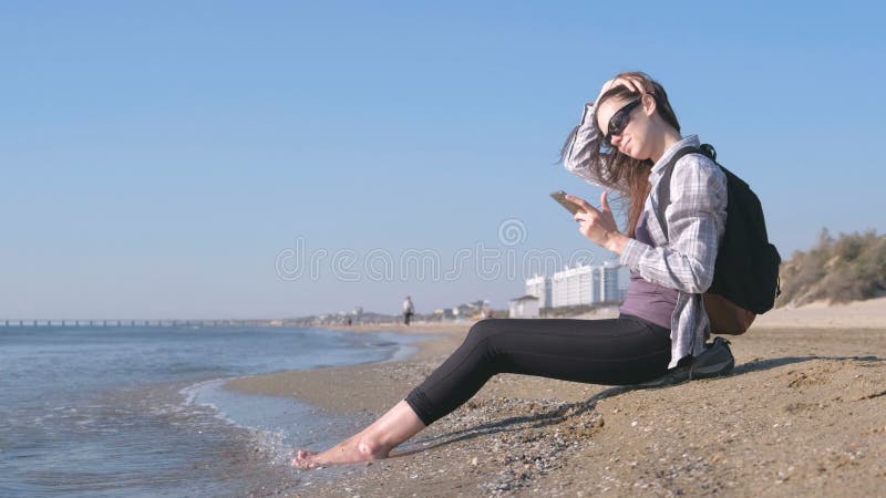 Menina do viajante com uma trouxa que senta-se em uma praia arenosa do mar e que datilografa uma mensagem no telefone celular