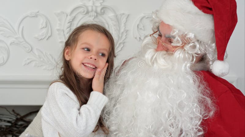 Menina doce no regaço de Santa Claus que diz lhe o que quer para o Natal fotografia de stock Menina doce no regaço de Santa Claus que diz lhe o que quer para o Natal fotografia de stock