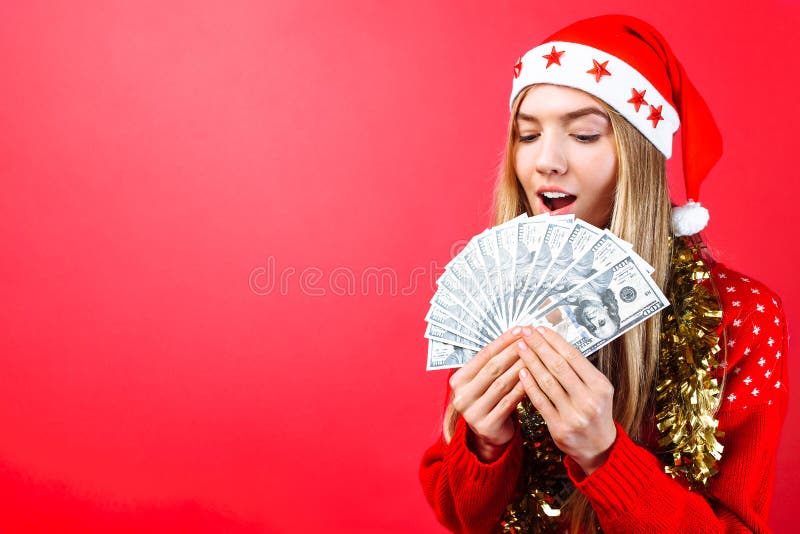 Menina emocional em uma camiseta e em um chapéu vermelhos de Santa Claus, na admiração que guarda o dinheiro em um fundo vermelho foto de stock Menina emocional em uma camiseta e em um chapéu vermelhos de Santa Claus, na admiração que guarda o dinheiro em um fundo vermelho foto de stock