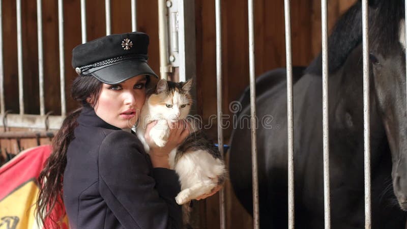 A menina encantador guarda um gato no estábulo em seus braços perto do cavalo preto