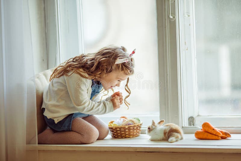 A menina encaracolado feliz bonita da criança está sentando-se no peitoril com seu coelho colorido pequeno do amigo, feriado da j fotos de stock A menina encaracolado feliz bonita da criança está sentando-se no peitoril com seu coelho colorido pequeno do amigo, feriado da j fotos de stock