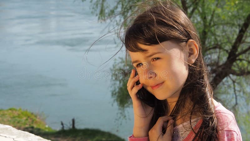 A menina está falando no telefone pela água Uma criança com um telefone em um dia ensolarado fora Menina com um telefone no rio