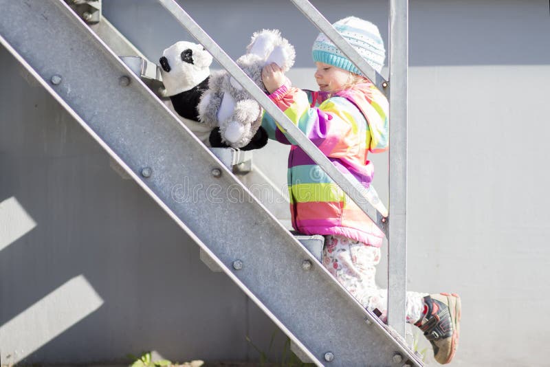 A menina está jogando na escadaria do metal com brinquedos do luxuoso O bebê no revestimento e no chapéu coloridos joga perto da imagem de stock A menina está jogando na escadaria do metal com brinquedos do luxuoso O bebê no revestimento e no chapéu coloridos joga perto da imagem de stock