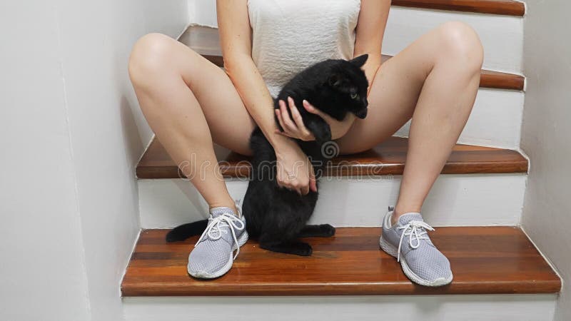 A menina está sentando-se nas escadas na casa entre seus pés está guardando um gato preto e está afagando-o foto de stock A menina está sentando-se nas escadas na casa entre seus pés está guardando um gato preto e está afagando-o foto de stock