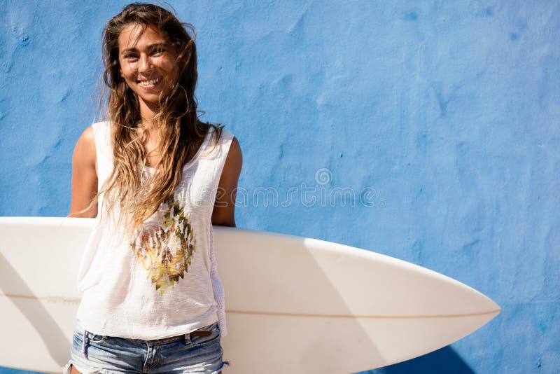 Menina feliz do surfista com a prancha na frente da parede azul foto de stock Menina feliz do surfista com a prancha na frente da parede azul foto de stock