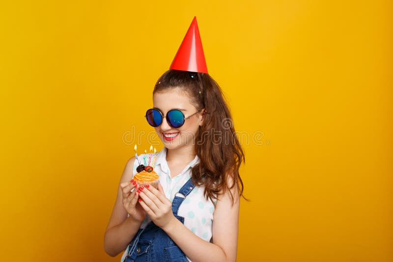 Menina feliz nos óculos de sol, sobre o fundo amarelo, realizando nas mãos um queque com velas imagens de stock Menina feliz nos óculos de sol, sobre o fundo amarelo, realizando nas mãos um queque com velas imagens de stock