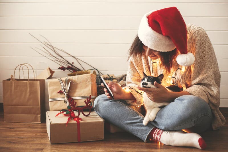 Menina feliz à moda no chapéu de Santa que olha a tela do telefone com o gato bonito em luzes de Natal no fundo dos presentes Mod imagem de stock royalty free Menina feliz à moda no chapéu de Santa que olha a tela do telefone com o gato bonito em luzes de Natal no fundo dos presentes Mod imagem de stock royalty free