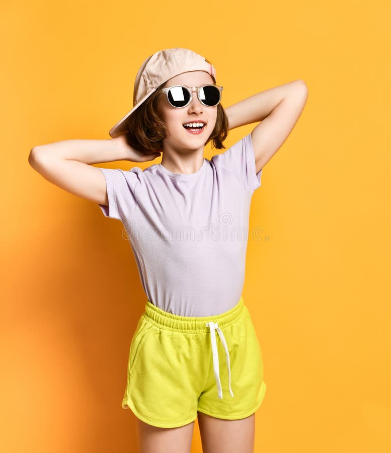 Menina fresca bonita do adolescente da forma nos óculos de sol e tampão, chapéu e short claros em um fundo amarelo foto de stock royalty free Menina fresca bonita do adolescente da forma nos óculos de sol e tampão, chapéu e short claros em um fundo amarelo foto de stock royalty free