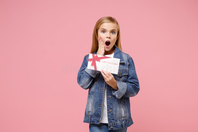 Shocked little blonde kid girl 12-13 years old wearing denim jacket posing  on pastel pink background. Childhood lifestyle concept. Mock up copy space. Hold gift certificate put hand on cheek. Shocked little blonde kid girl 12-13 years old wearing denim jacket posing  on pastel pink background. Childhood lifestyle concept. Mock up copy space. Hold gift certificate put hand on cheek
