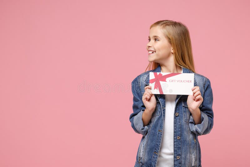 Smiling little blonde kid girl 12-13 years old in denim jacket posing isolated on pastel pink wall background. Childhood lifestyle concept. Mock up copy space. Hold gift certificate, looking aside. Smiling little blonde kid girl 12-13 years old in denim jacket posing isolated on pastel pink wall background. Childhood lifestyle concept. Mock up copy space. Hold gift certificate, looking aside