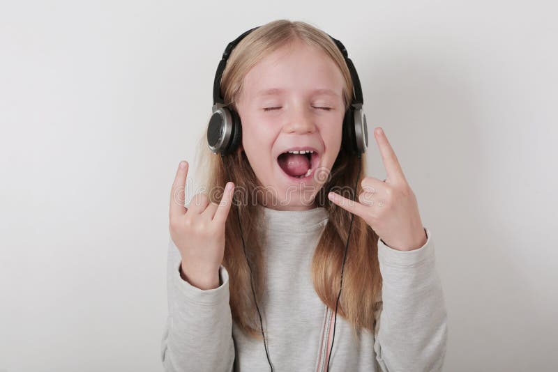 Menina loura com música de escuta e canto dos fones de ouvido Menina bonito que faz um sinal do rocha-n-rolo imagens de stock Menina loura com música de escuta e canto dos fones de ouvido Menina bonito que faz um sinal do rocha-n-rolo imagens de stock