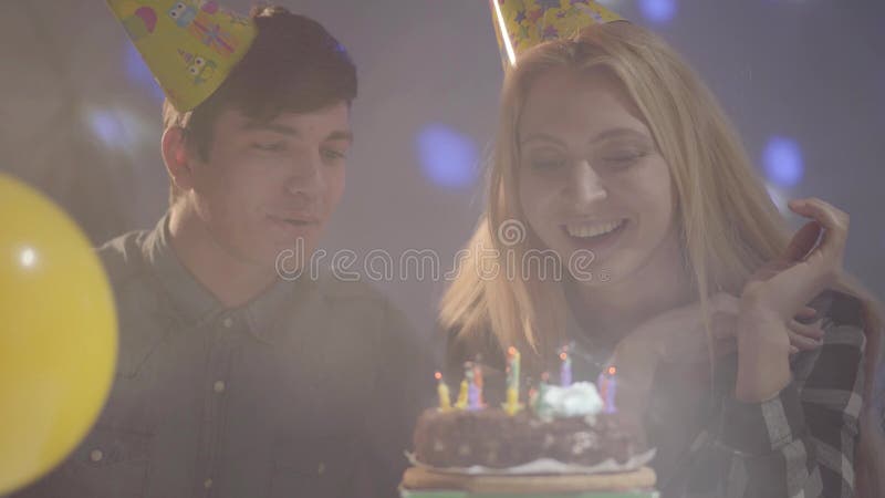 Menina loura de sorriso bonito no chapéu do aniversário que funde para fora velas no bolo, homem novo que senta-se próximo A mulh