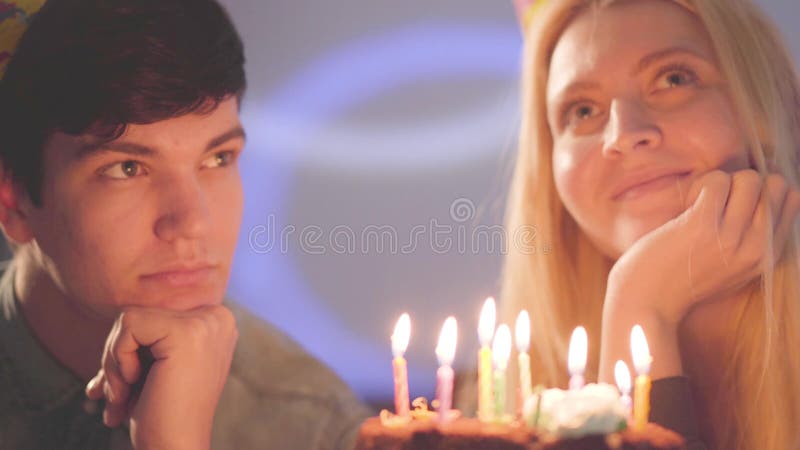 Menina loura do retrato do close-up e um homem nos chapéus do aniversário que sentam-se na frente do bolo com velas A mulher tem