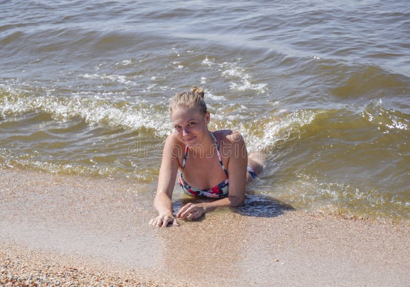 A menina loura em um biquini que encontra-se na praia e as ondas espirram nela Jovem mulher bonita em um biquini colorido no fund fotografia de stock A menina loura em um biquini que encontra-se na praia e as ondas espirram nela Jovem mulher bonita em um biquini colorido no fund fotografia de stock
