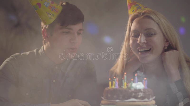 A menina loura no chapéu do aniversário que funde para fora velas no bolo, homem novo que senta-se próximo A mulher tem o anivers