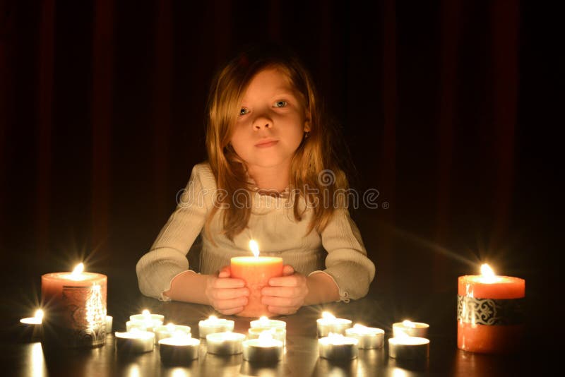 A menina loura pequena bonito está guardando uma vela ardente, lotes das velas é em torno dela sobre o fundo escuro foto de stock A menina loura pequena bonito está guardando uma vela ardente, lotes das velas é em torno dela sobre o fundo escuro foto de stock