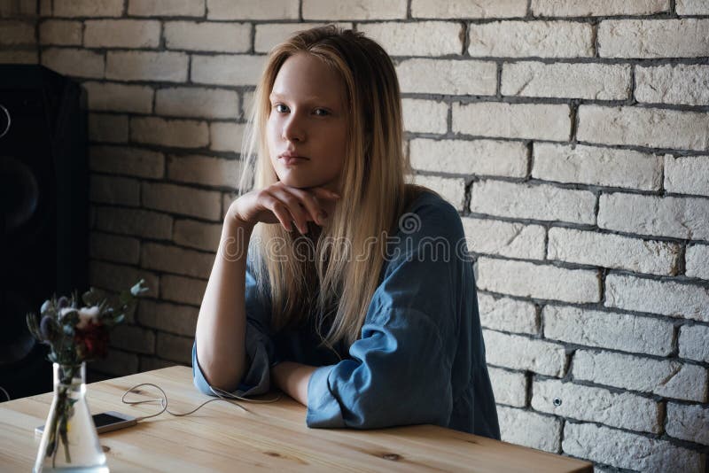 A menina loura senta-se com fones de ouvido e olha-se pensativamente ausente, com sua mão em seu queixo imagem de stock royalty free A menina loura senta-se com fones de ouvido e olha-se pensativamente ausente, com sua mão em seu queixo imagem de stock royalty free