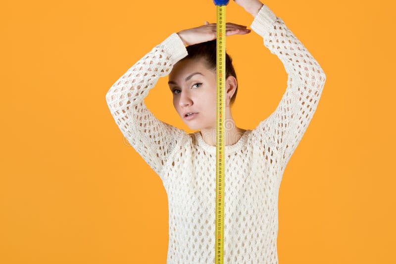 Girl measures her height, holding a measuring tape nearby and looks at the camera, isolated on orange-yellow. Girl measures her height, holding a measuring tape nearby and looks at the camera, isolated on orange-yellow