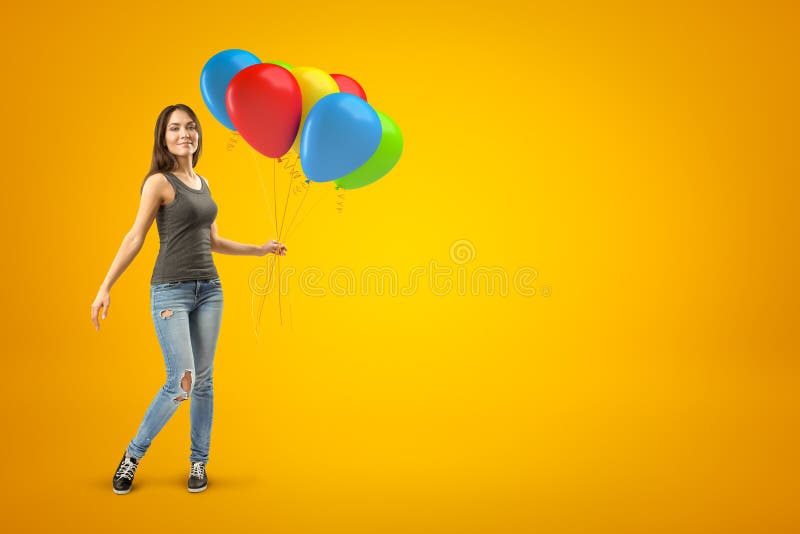 Menina moreno nova que veste cal?as de brim ocasionais e o t-shirt que levam bal?es coloridos no fundo amarelo imagens de stock Menina moreno nova que veste cal?as de brim ocasionais e o t-shirt que levam bal?es coloridos no fundo amarelo imagens de stock