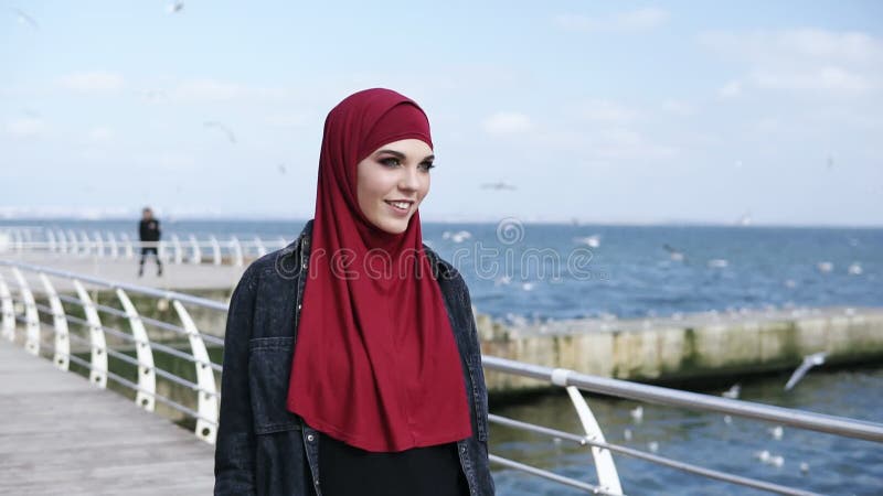 A menina muçulmana atrativa nova aprecia ter uma caminhada perto do lado de mar com as gaivotas que voam ao redor no fundo