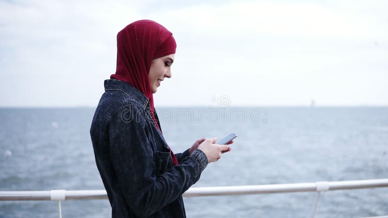 A menina muçulmana atrativa nova com hijab em sua cabeça é de enrolamento e de datilografia algo em seu smartphone ao andar