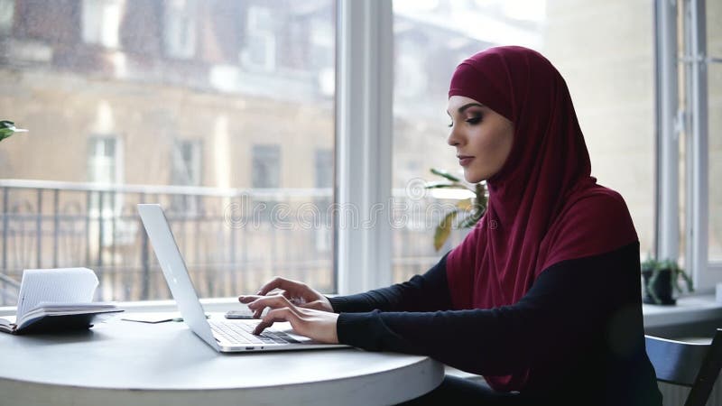 A menina muçulmana atrativa nova com o hijab que cobre sua cabeça está datilografando algo em seu portátil ao sentar-se em algum