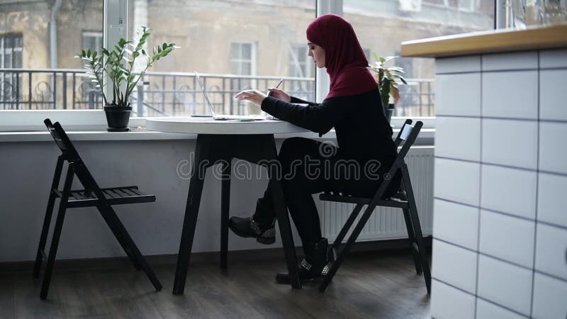 A menina muçulmana bonita com hijab em sua cabeça, sentando-se perto da janela, está datilografando abaixo de algo no teclado del